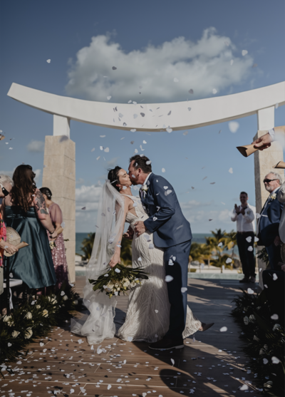 A bride and groom share a kiss under a shower of white confetti at their tropical beach wedding, celebrating their love in a stunning oceanfront ceremony.