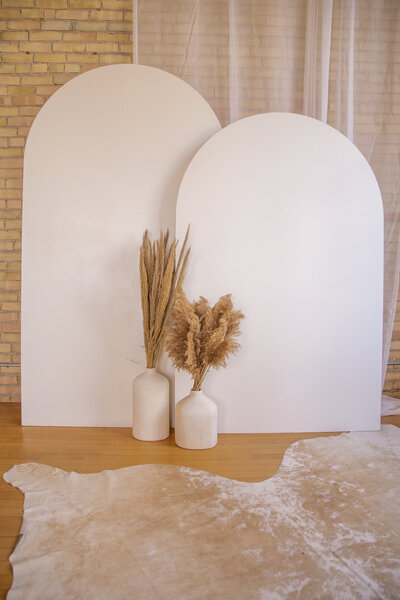 Pampas grass in two stone vases on the ground, sitting in front of two large white wooden arch backdrops.