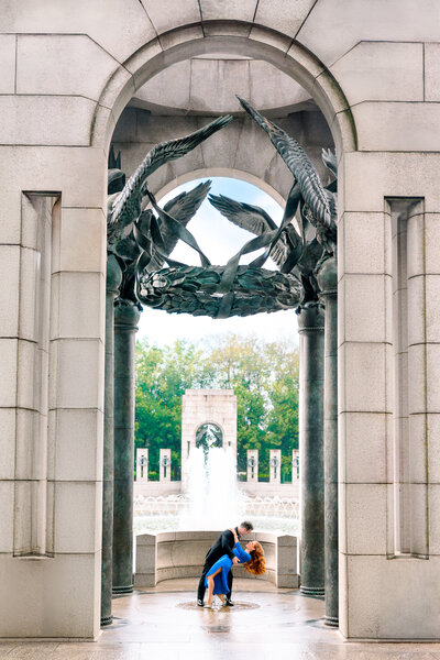 A couple dances gracefully beneath a large stone arch adorned with metal sculptures of birds in flight. They are framed by the archway, with a fountain and trees visible in the background under a clear sky.