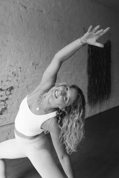 Black and white photo of woman with long, blonde, curly hair smiles and stretches with her arm reacher overhead wearing yoga close.