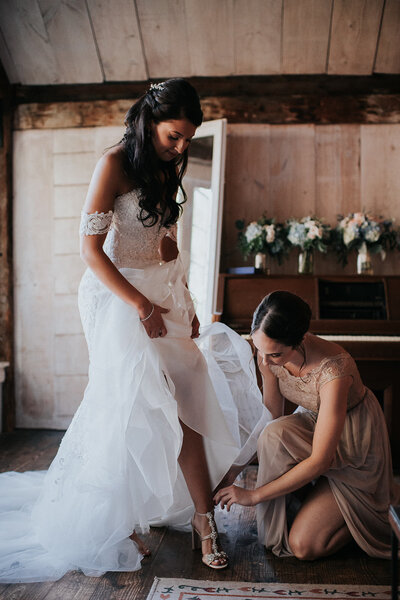 Bridesmaid helps bride buckle her shoes while getting ready on her wedding day