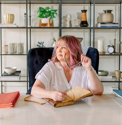 Woman with white skin and pink hair hold glasses and writing in journal, looking out the window near desk.