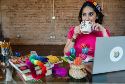 The world of creativity as the founder of our art workshops sits at her glass desk. Discover the artistic tableau featuring a personal laptop, handmade decorations, an assortment of brushes, books with imaginative covers, and lively pompoms.