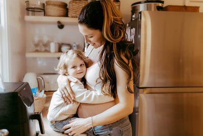A woman hugs a child in a kitchen. The child is smiling while the woman looks down at them affectionately. They stand next to a stainless steel refrigerator.