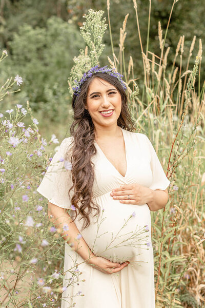 Woman in v-neck cream dress with long brown hair in loose curls coming over one shoulder. She is holding her pregnant belly and smiling towards the camera. She is standing in a field of tall wildflowers