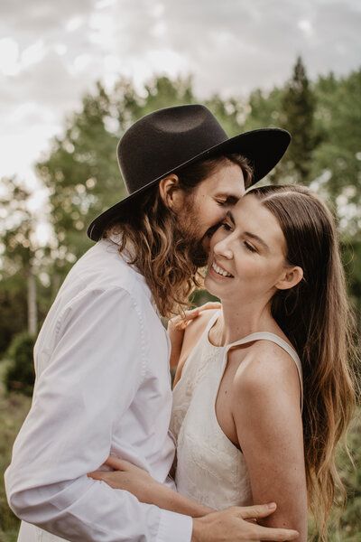 engaged couple embracing each other as the man with long hair, a bread and a black hat kisses the woman's cheek and she smiles