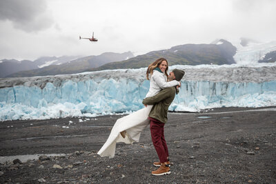 A bride and groom embrace joyfully on a rocky glacier landscape in Alaska, with a helicopter flying overhead and towering ice formations in the background. The bride’s white dress flows in the wind as the groom lifts her, capturing the excitement of a helicopter elopement in the Alaskan wilderness.