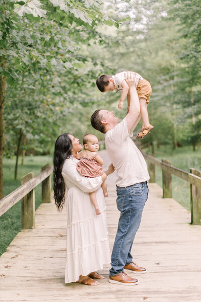 A joyful family of four sharing a playful moment on a wooden bridge surrounded by trees — Raleigh portrait photography.