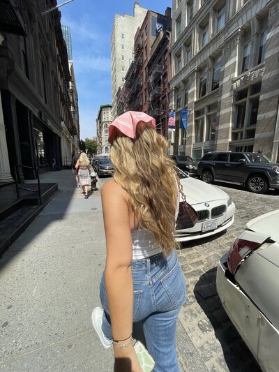 Woman with long blonde hair and a pink cap walks along a sunny city street. She's wearing jeans and a white top, surrounded by parked cars and tall buildings.