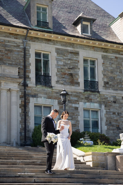 Aldrich Mansion Wedding Photos | Kelsey Sheehan Photography Timeless Rhode Island Weddings | A bride and groom stand on steps before a grand stone building, with the bride reading from a card. Both show a tender, joyful demeanor.