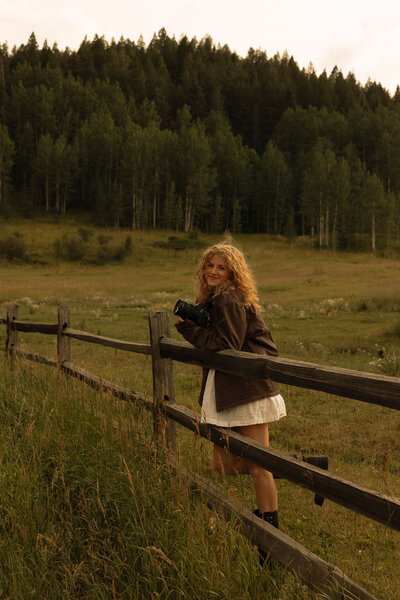 A woman with long blonde hair leans on a wooden fence in a serene, grassy field surrounded by trees in colorado, holding a camera and exuding a timeless, nostalgic vibe.