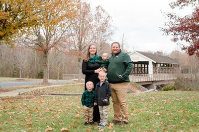A family of five, including two adults, two young boys, and a baby, stand outdoors in a grassy area with autumn trees and a covered bridge in the background.
