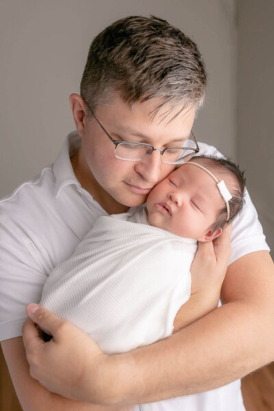 Dad with glasses and light brown hair is holding his newborn baby girl up close by his face and is resting his cheek on her cheek. Baby girl is sleeping and swaddled in a white blanket and wearing a minimalist white bow on her forehead. Dad has his eyes closed and is soaking up the moment with his new baby.