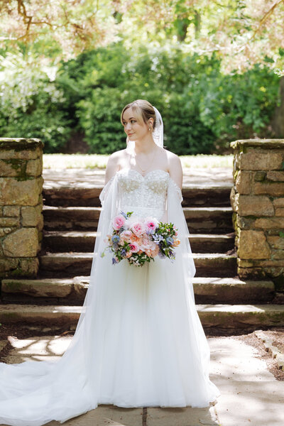 A radiant bride stands in soft sunlight at Ridley Creek Parque Mansion, holding a lush bouquet of pastel flowers. She wears an elegant off-the-shoulder wedding gown with delicate lace detailing and a flowing train. The natural stone steps and greenery create a romantic, timeless setting.