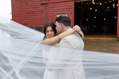 Bride smiles over her shoulder as the groom nuzzles into her head, veil flowing dramatically in the wind
