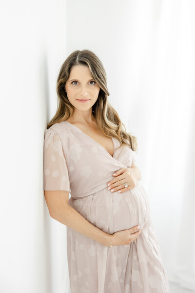 Pregnant woman embraces her belly in a blush colored dress during in-studio maternity portrait session