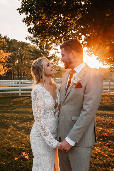 bride and groom hold hands as the sun sets behind them in a field with white horse fencing