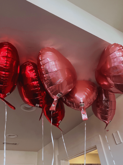 A cluster of red heart-shaped foil balloons floating under a ceiling. The balloons are tied together with white ribbons.