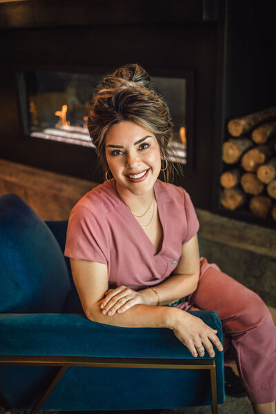 woman sitting in a blue chair in front of a fireplace wearing pink scrubs and smiling with her hair in a bun