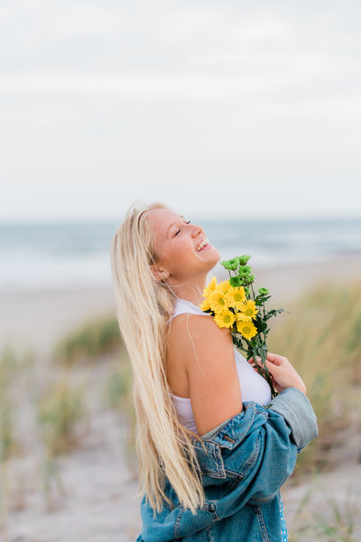 Picture of a high school senior girl laughing while holding flowers. She is on the beach wearing a denim jacket. Captured by Nicole Marie Photography in Ocean City, New Jersey.