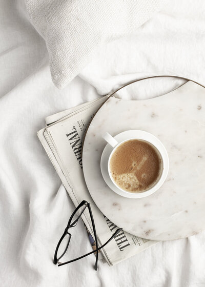 A cup of coffee, newspaper, and a pair of glasses on top of a white bed sheet
