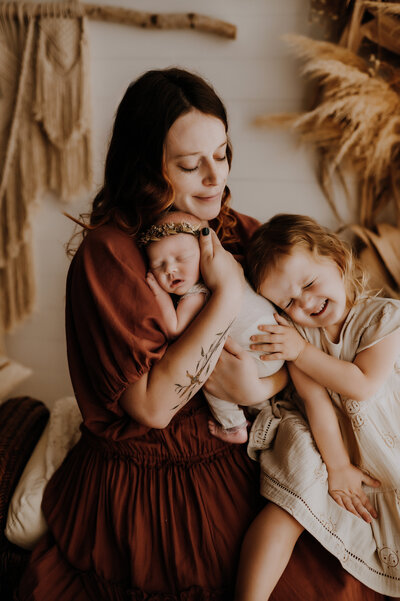 A mom holding her newborn baby to her chest while her other toddler hugs both of them and smiles in a fort worth, texas photography studio.
