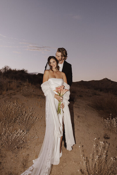 Couple embracing during their elopement at sunset in Joshua Tree, surrounded by towering rock formations and iconic Joshua trees with a warm, golden sky in the background.