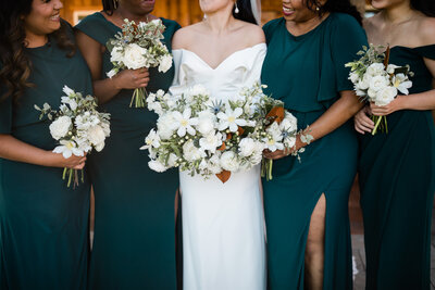 bride stands with bridesmaids in hunter green dresses holding white & green flowers with magnolia leaves and thistles