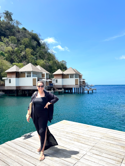 Woman in black walking on dock over blue waters with over-water bungalows behind her