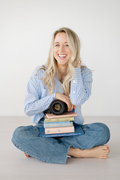 Happy photographer sitting on ground, holding her camera and storybooks, joyfully laughing.