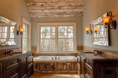 An interior shot of a rustic but rich bathroom. Everything is wood paneled and a large porcelain soaking tub sits beneath large windows.