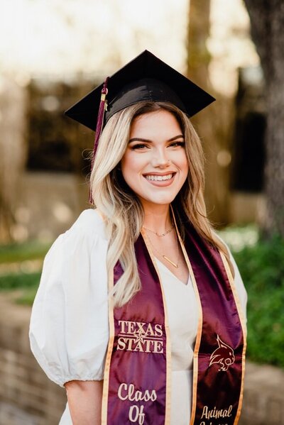 A smiling graduate wearing a cap and maroon Texas State stole stands outdoors, exuding pride and accomplishment. Bright, natural light enhances the scene.