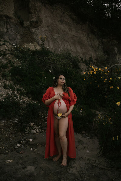 Maternity photo of woman standing out in a wooded area in a long red wrap with her belly exposed, holding a yellow flower to her stomach, captured by Morgan Ashley Photography in Milwaukee, WI