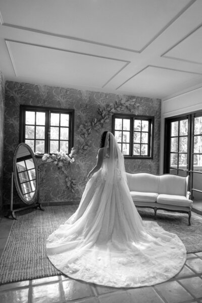 Monochromatic, dramatic image of a bride seen from behind, sitting in her long wedding dress and veil in a luxury getting ready room with dark paneled walls, a couch, and a standing mirror. 