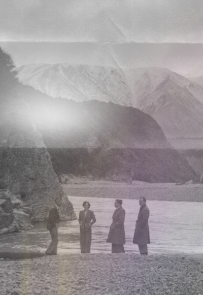 Retro black and white photo of four people stand on the beach with mountains in the background.