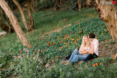 Engaged couple cozy up as they sit together under a tree surrounded by green and orange flowers at Gum Grove Park