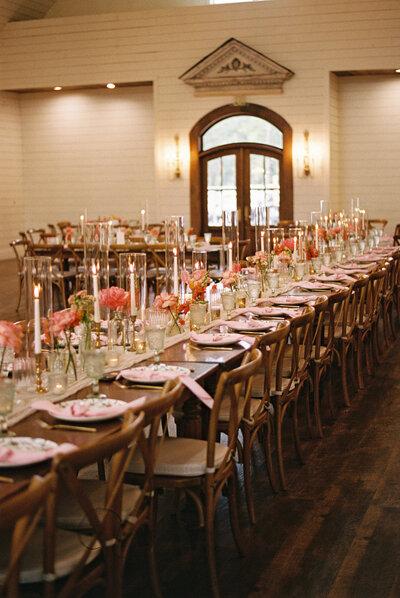 A long banquet table, elegantly set with pink accents, extends through a light-colored room, likely a reception hall.