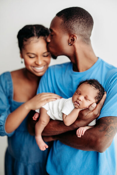 Cute African American newborn photographed posed in his dad's arms while dad kisses mom on the temple and mom smiles down at baby in Jen Howell Photography's Memphis newborn photography studio.