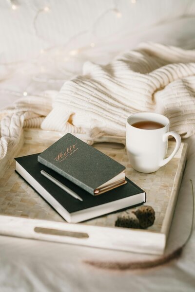 A stack of notebooks, pen, and cup of coffee sit on a tray table on a bed.
