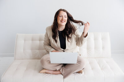 This is Mariah sitting on a white couch with her laptop and legs crossed. She is smiling and playing with her hair. 