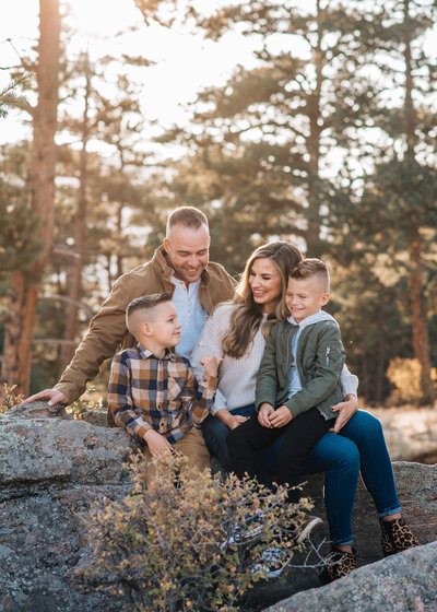 denver family photographers captures young family in the woods sitting together on a rock while the sun sets in the distance that shines through the trees of the woods