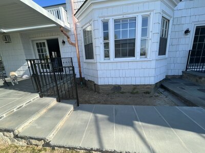 A home entrance with a dull patio, bare soil, and no landscaping. The white house has black railings, and the area appears unfinished.