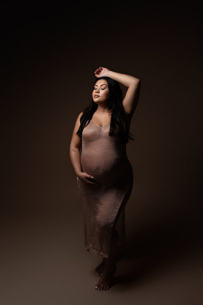 Professional Austin maternity portrait of pregnant woman in form-fitting nude dress with dramatic studio lighting and classic pose