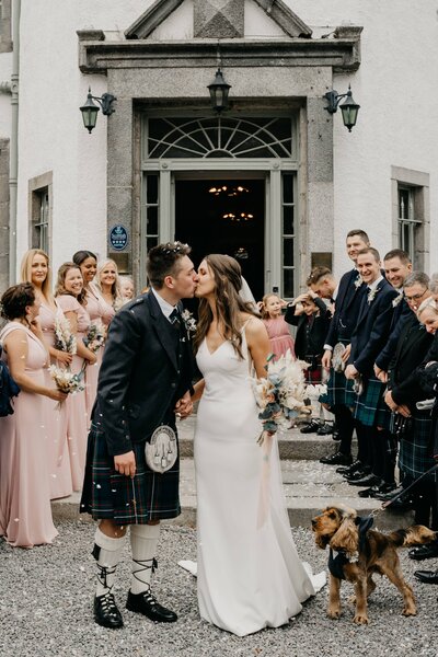 A wide photograph of a bride and groom kissing outside Banchory Lodge Hotel. Their guests stand in rows either side of them after their wedding ceremony and throw confetti high in the air.