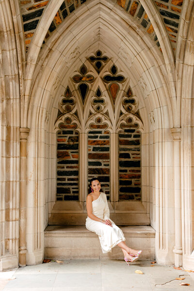 Duke University graduate in white dress and heels sitting in the side arch of Duke Chapel.