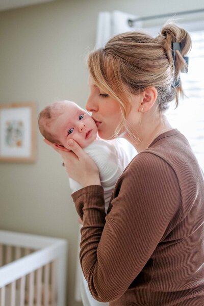 Sweet lifestyle moment of a mother kissing her baby in a cozy nursery, with the baby gazing softly into the camera.