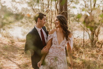 A bride in a boho lace gown and her partner walk through a wooded area during their intimate forest elopement in Spring, Texas, sharing a laughing moment surrounded by golden light.