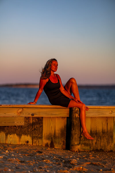 Blonde woman sits on a wooden ocean dock at sunset wearing a black yoga onesie looking into the distance.