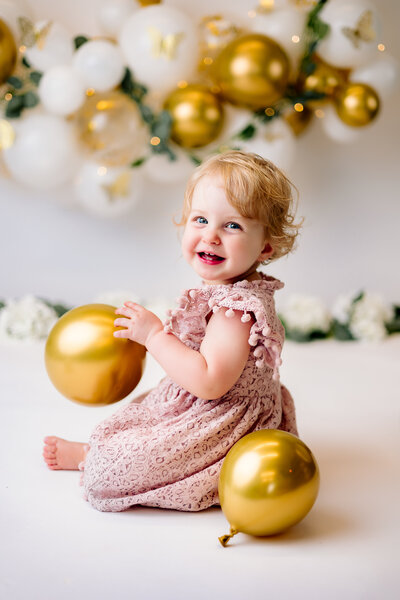 Baby girl turns back with a huge smile on her face,  playfully interacting  with a gold balloon during her milestone session