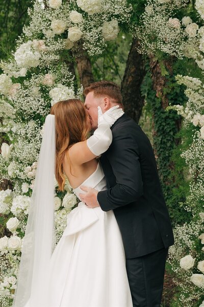 bride and groom saying vows under white floral arch during wedding ceremony 